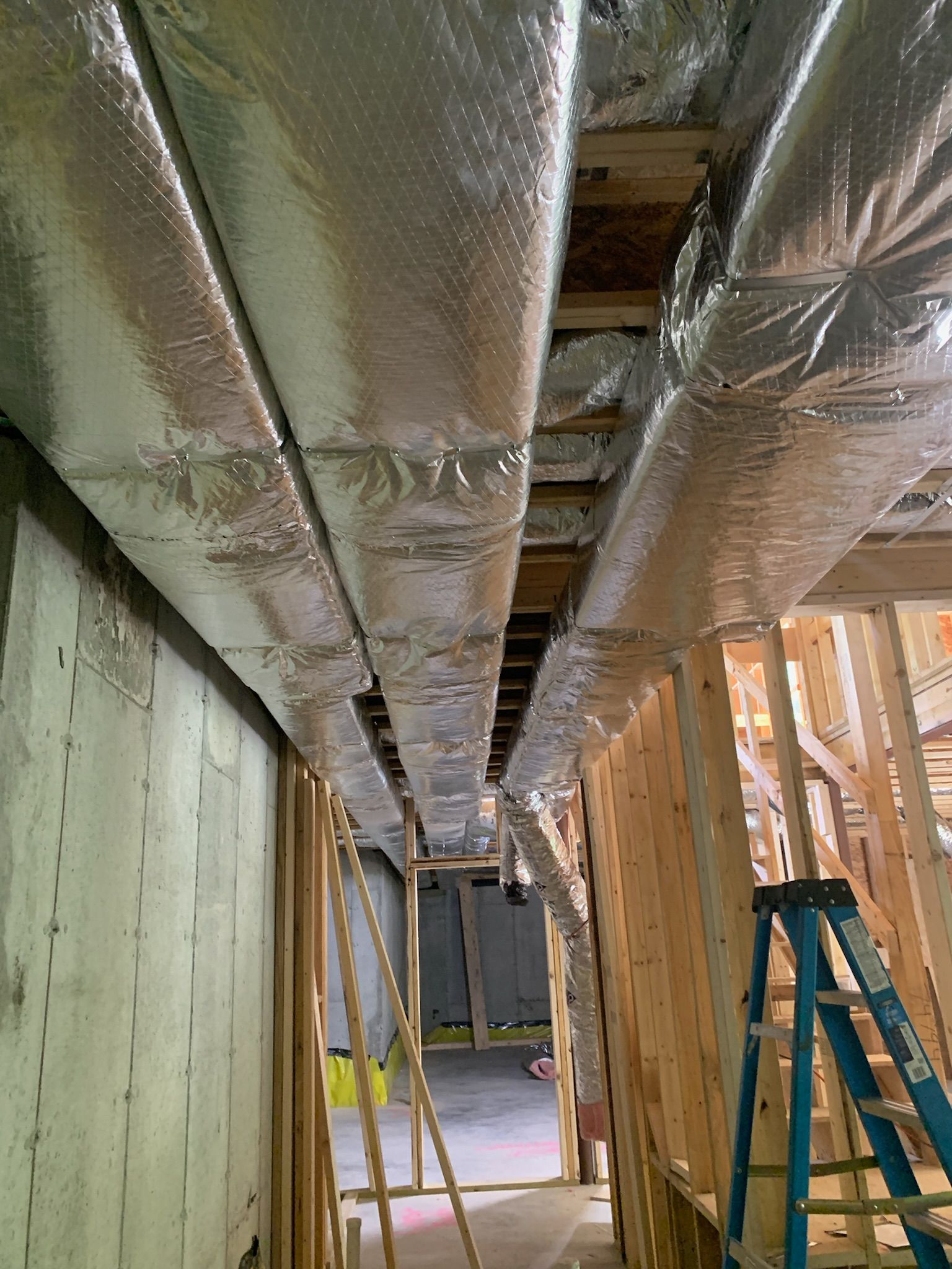 Ductwork wrapped in silver insulation runs across a partially constructed hallway with wooden framing and a blue ladder.
