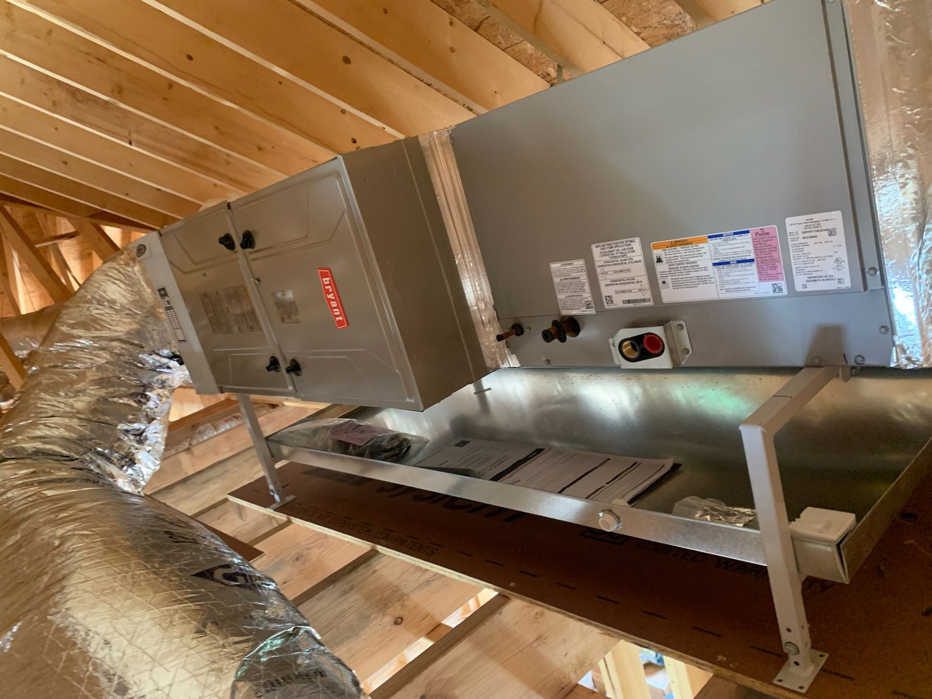 HVAC unit in an attic, silver metal, mounted on supports. Silver ductwork on the left, wooden rafters visible.