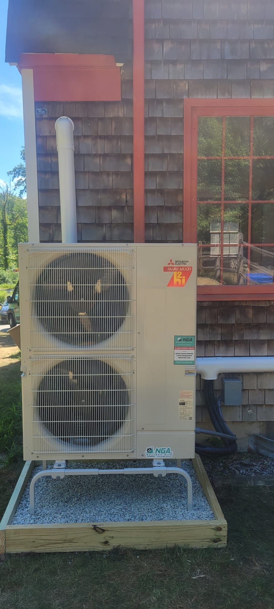An air conditioning unit on a gravel-filled wooden frame is mounted on the side of a brown shingled house.