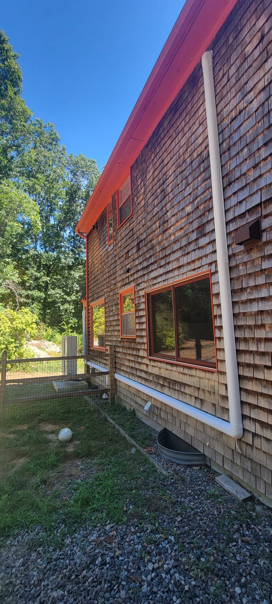 Side of a two-story wooden shingle building with a red roof, windows, and white drainpipe, on a sunny day.