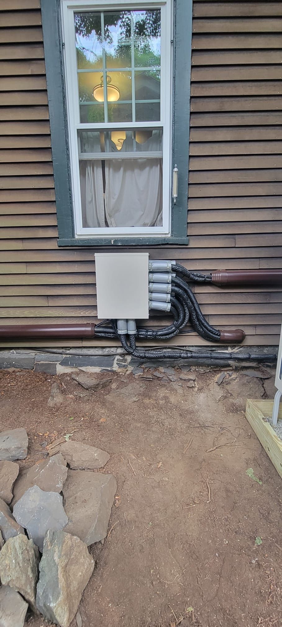 Exterior wall with window above electrical box and exposed conduit. Ground is dirt with rocks.