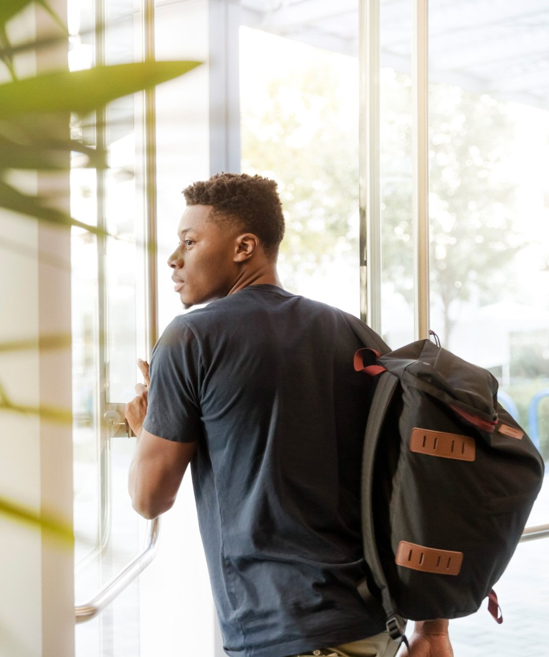 Man exiting a building, holding a backpack. He looks back at the entrance, standing in front of glass doors.