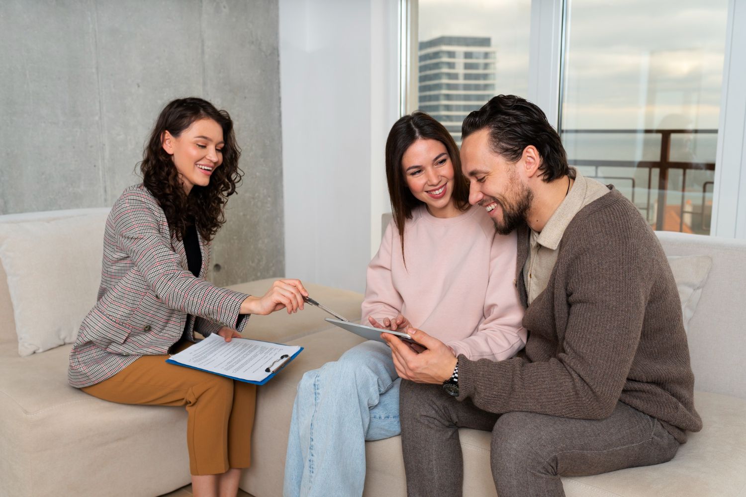 Three people on a couch discussing a tablet, with a woman holding a clipboard nearby