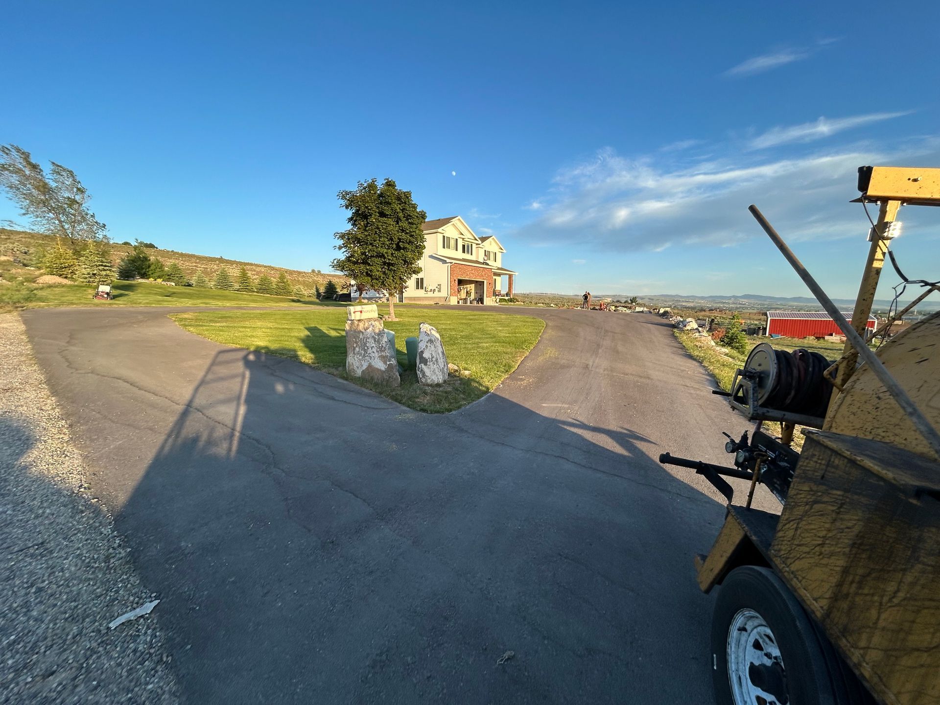 A paved driveway splits into two paths in front of a house at sunset, with a piece of construction equipment in the foreground.