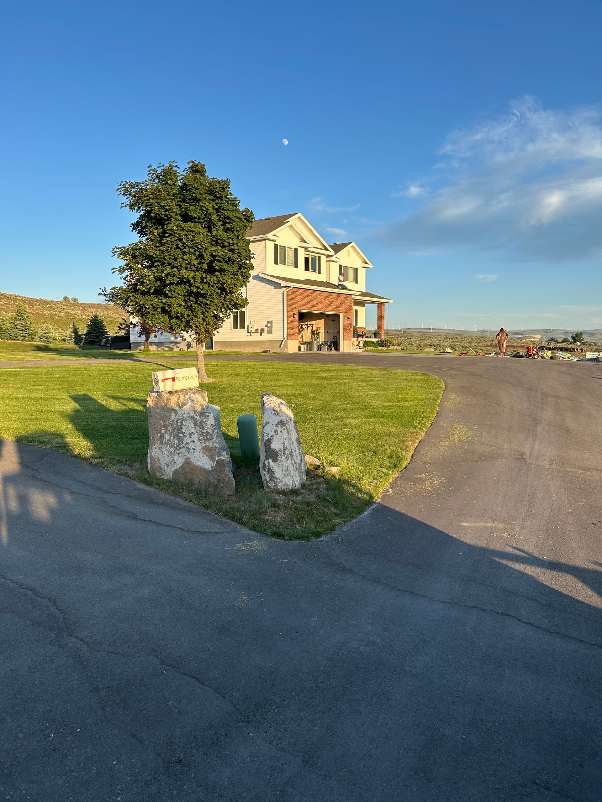 A two-story beige house with a brick entrance stands on a grassy lot under a clear blue sky, viewed from a driveway.