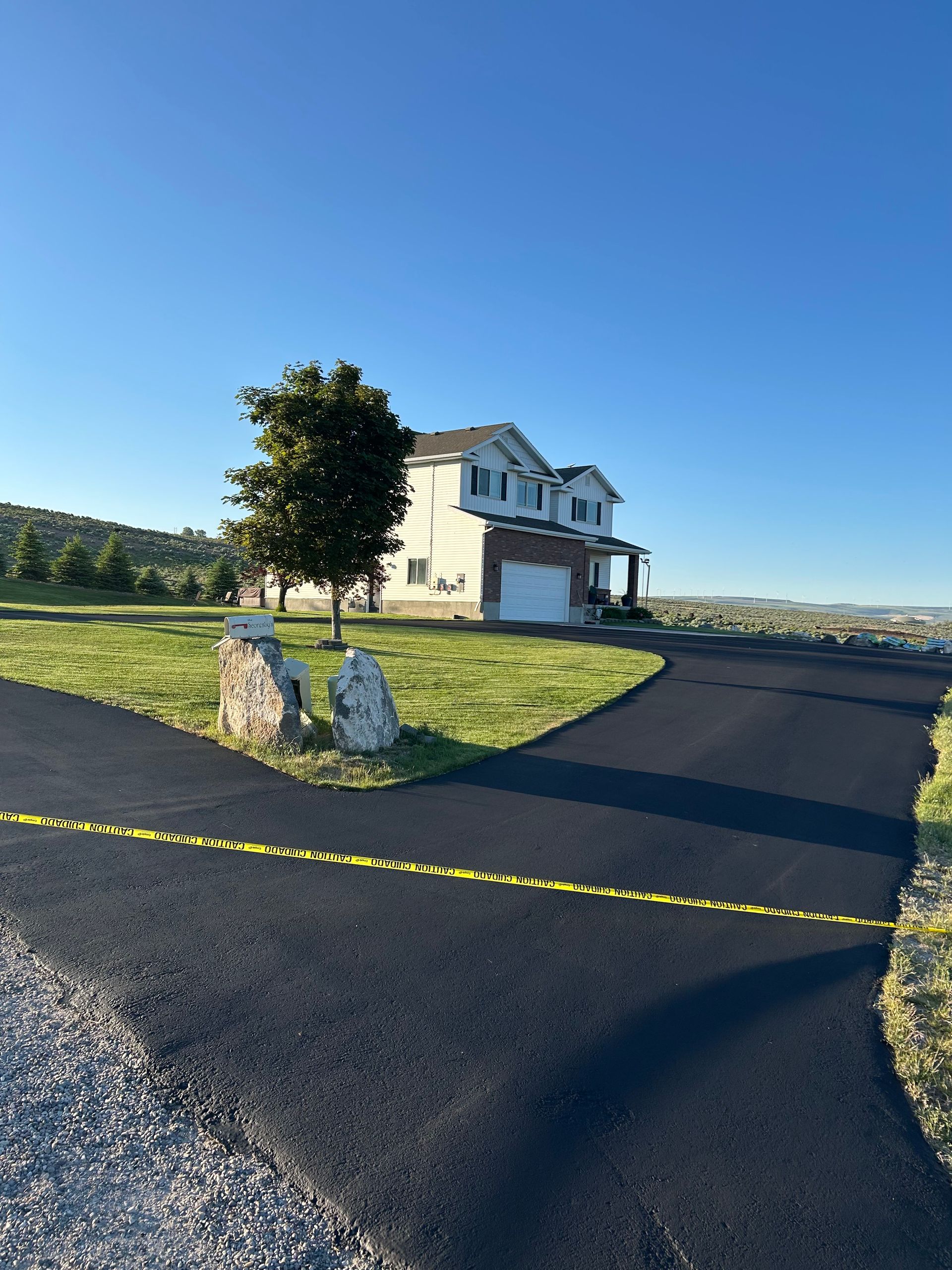 A two-story light-colored house with a dark roof behind a freshly paved asphalt driveway marked with yellow caution tape.