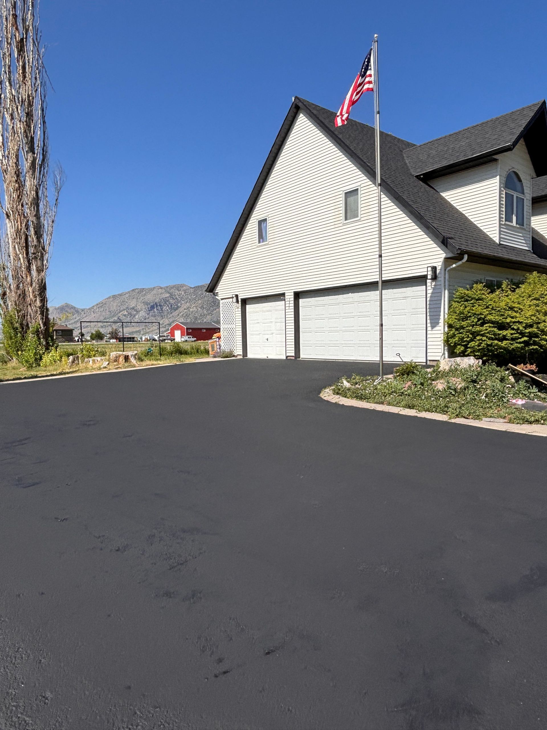A freshly paved asphalt driveway leading to a two-car garage of a white house with a mountain backdrop and an American flag.