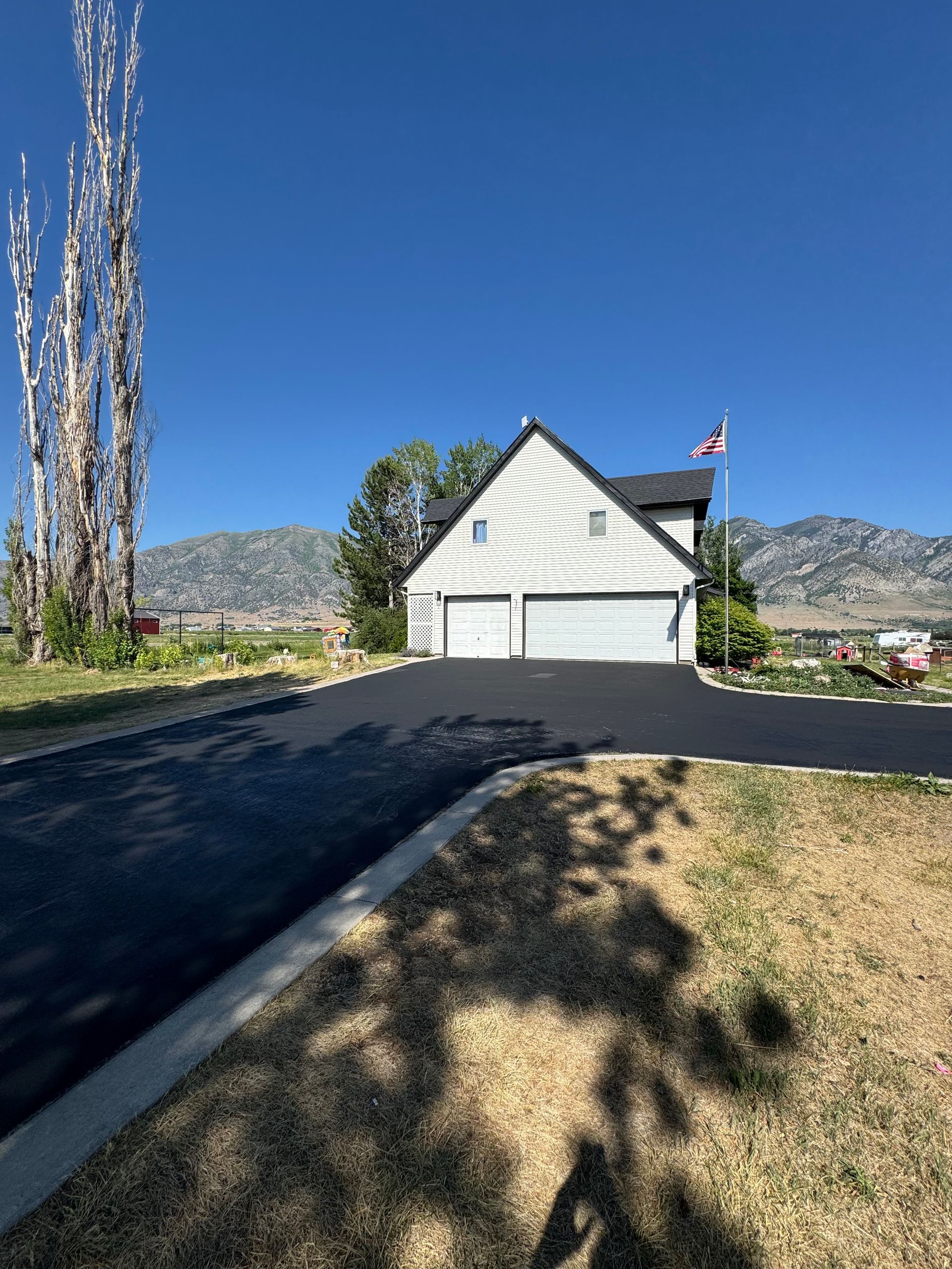 A white house with a garage and a new asphalt driveway, set against a backdrop of mountains under a clear blue sky.