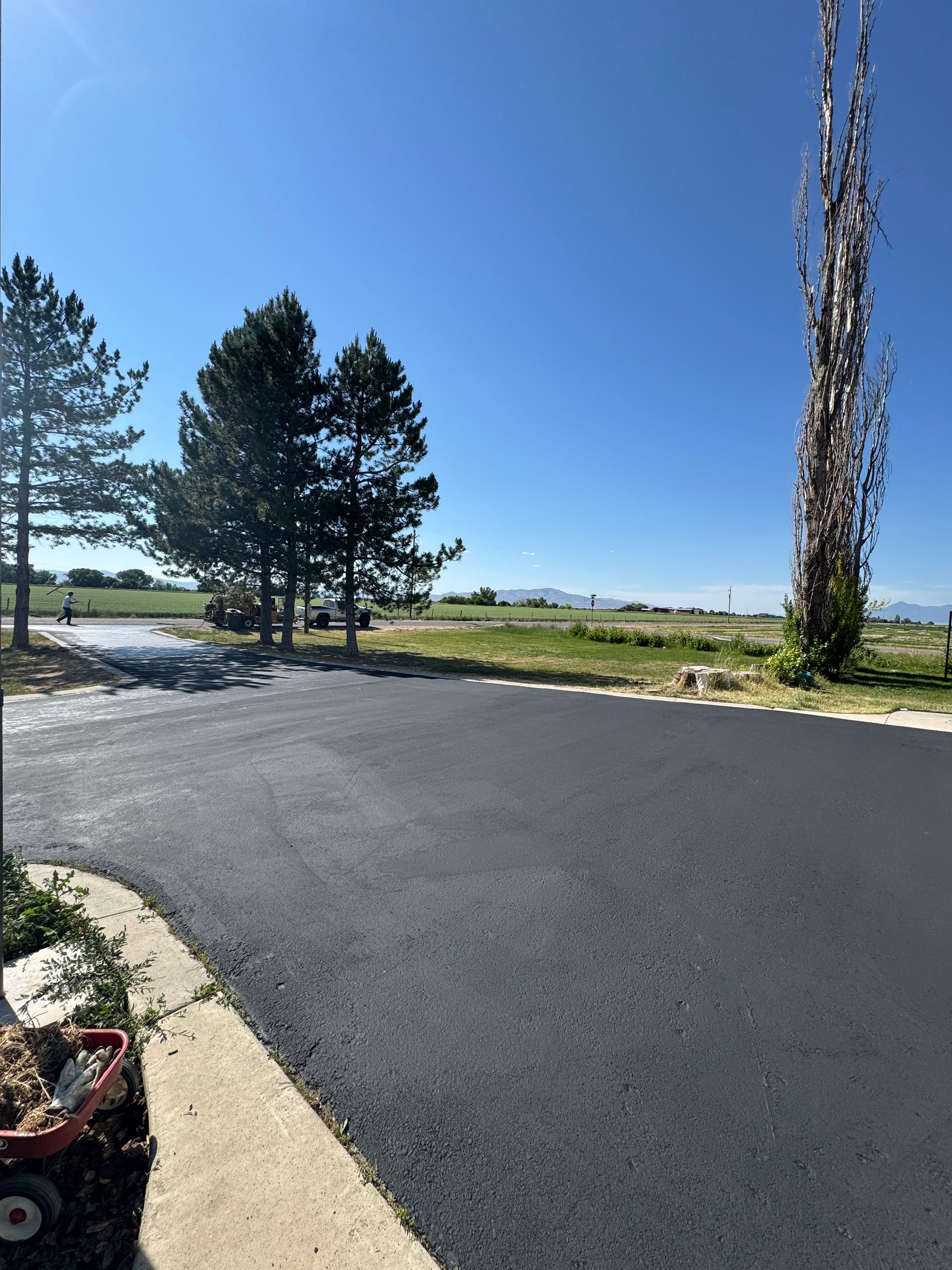 Freshly paved asphalt driveway leading toward a grassy field and distant hills under a clear blue sky.