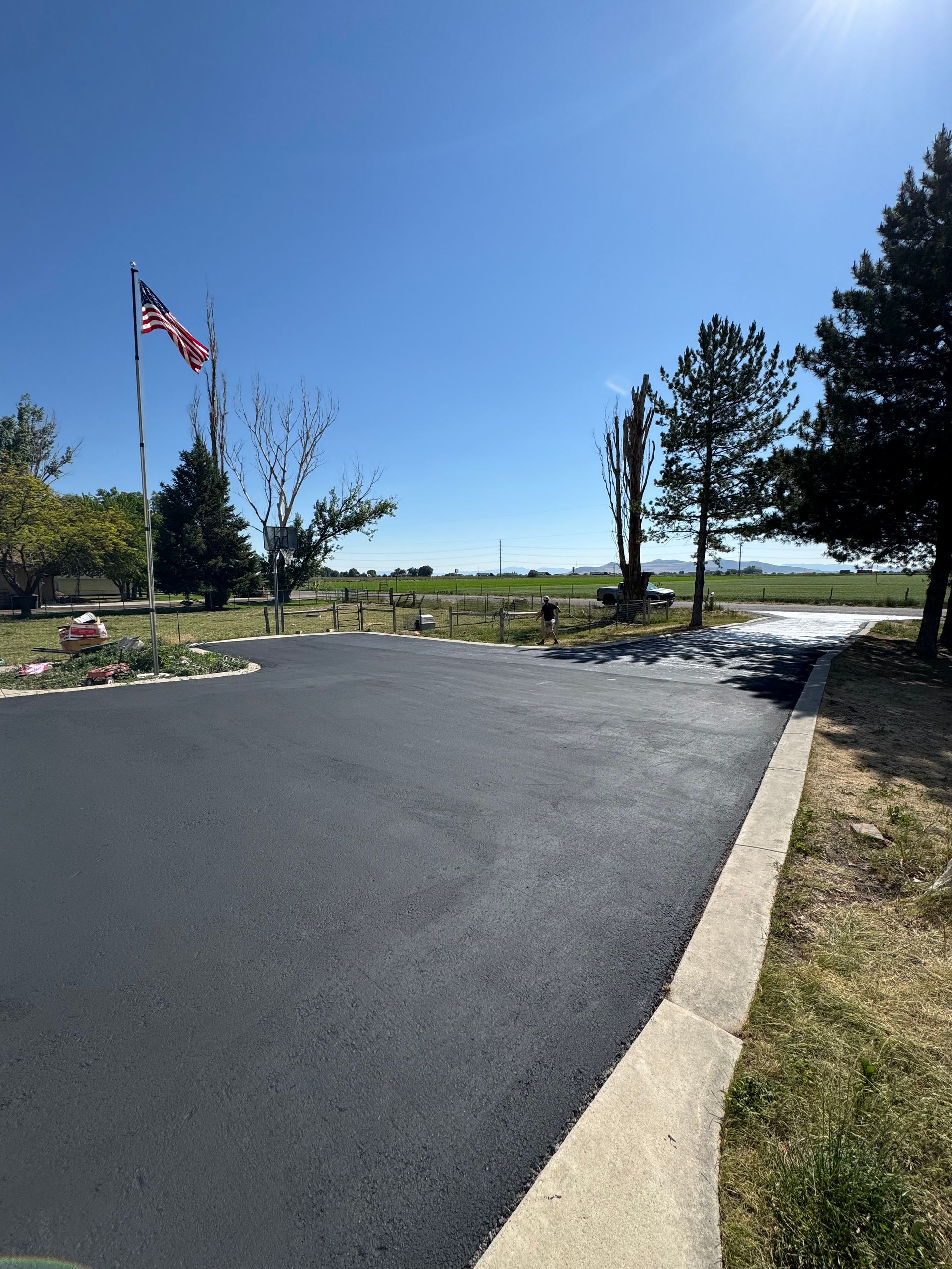 A freshly paved black asphalt driveway leads toward a grassy field under a clear blue sky, with a US flag on a tall pole.