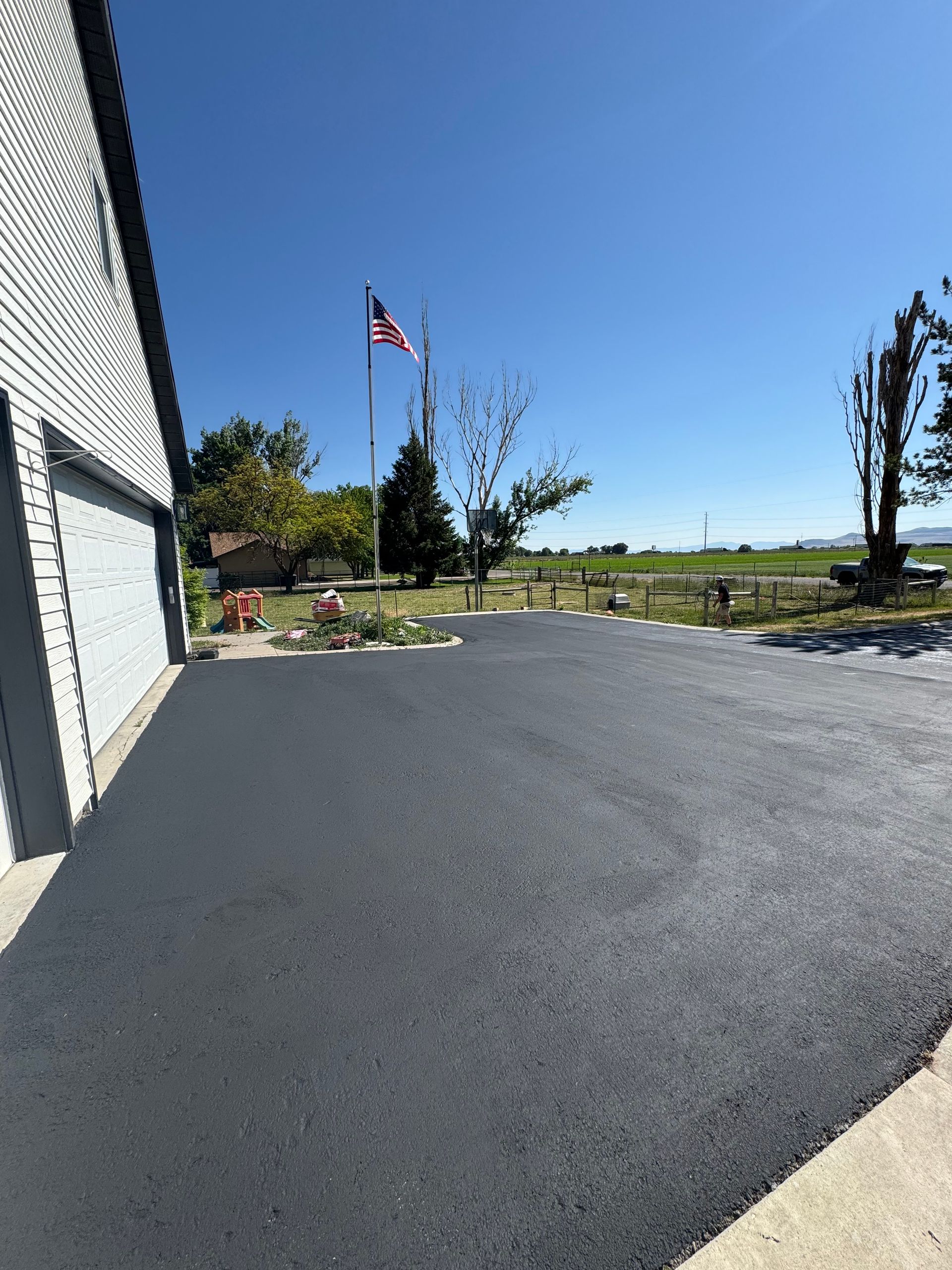 A freshly paved asphalt driveway leading to a house, with an American flag flying in the background under a blue sky.