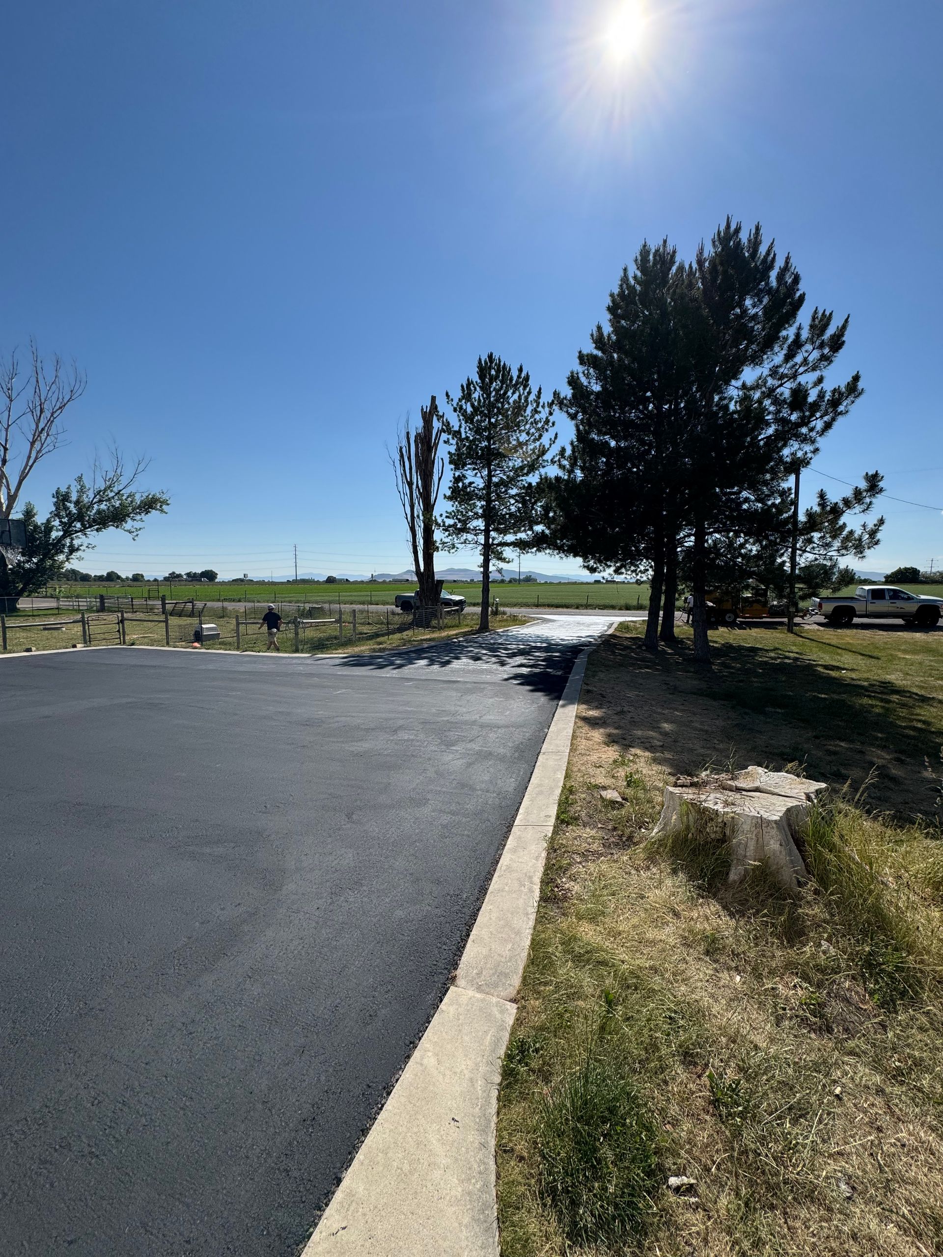 A newly paved black asphalt road leads toward a field and trees under a clear, bright blue sky with the sun shining.