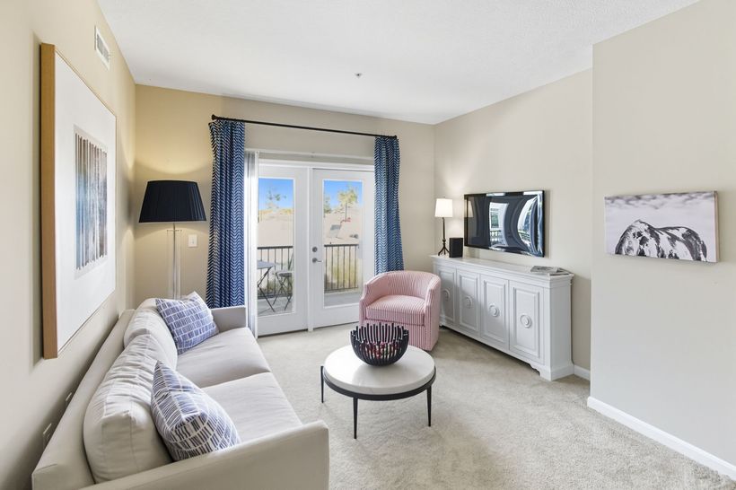 A bright, modern living room featuring a cream sofa, a coffee table, a dark armchair, and a glass door leading outside.