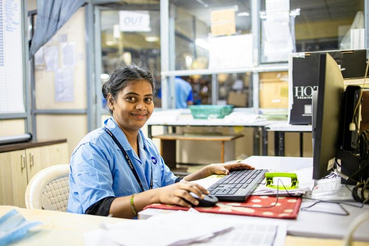 A smiling office worker in a blue uniform sits at a desk, typing on a computer keyboard in a bright, modern workspace.