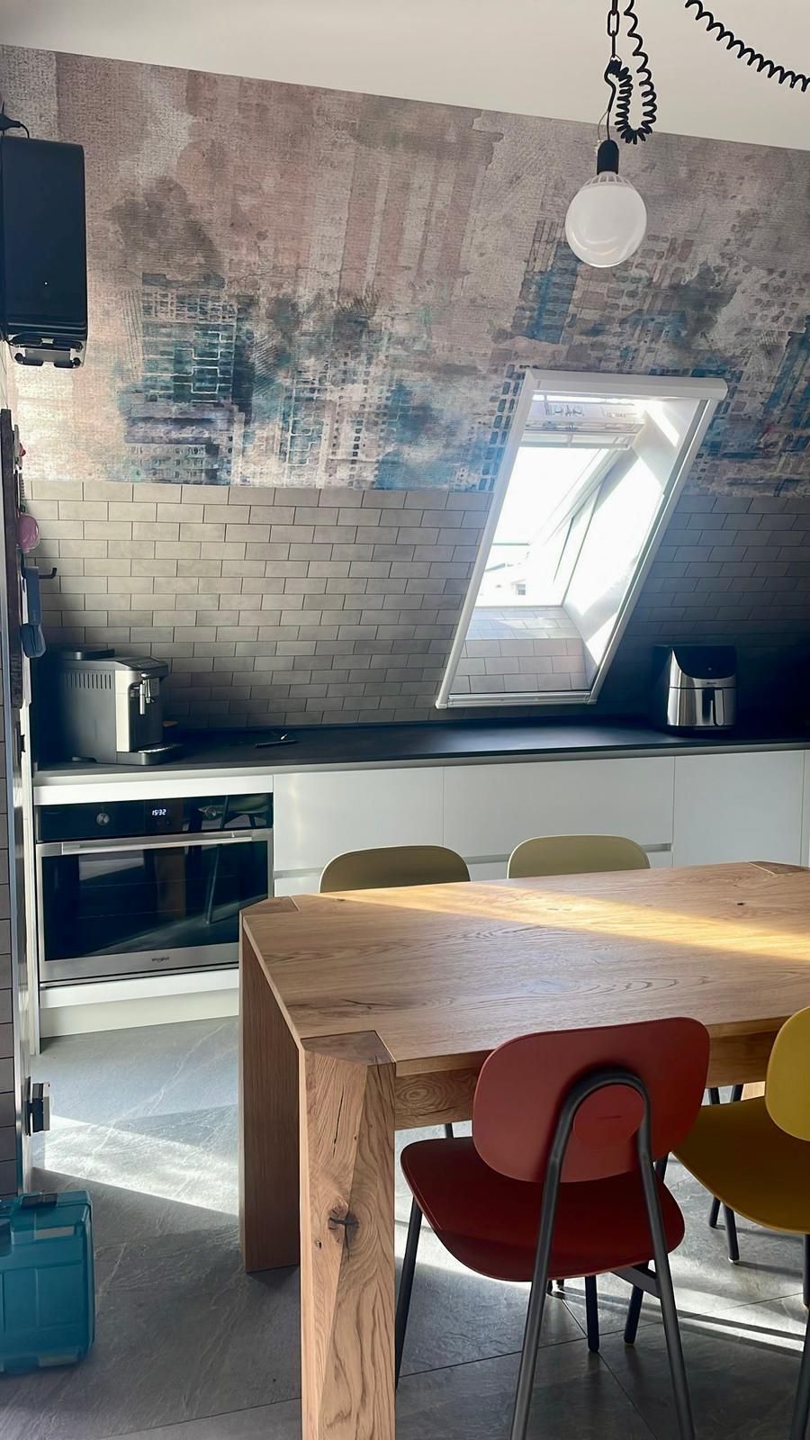 Cucina con tavolo in legno e sedie colorate, lucernario, parete in mattoni ed elettrodomestici.