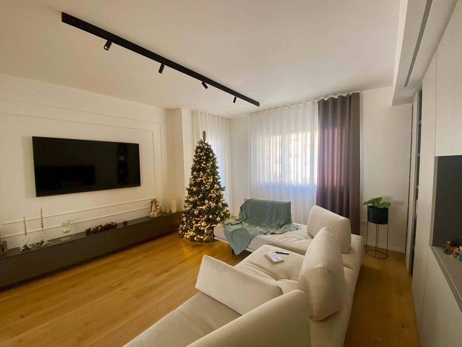 Soggiorno con albero di Natale, grande TV, divano componibile bianco e pavimento in legno.