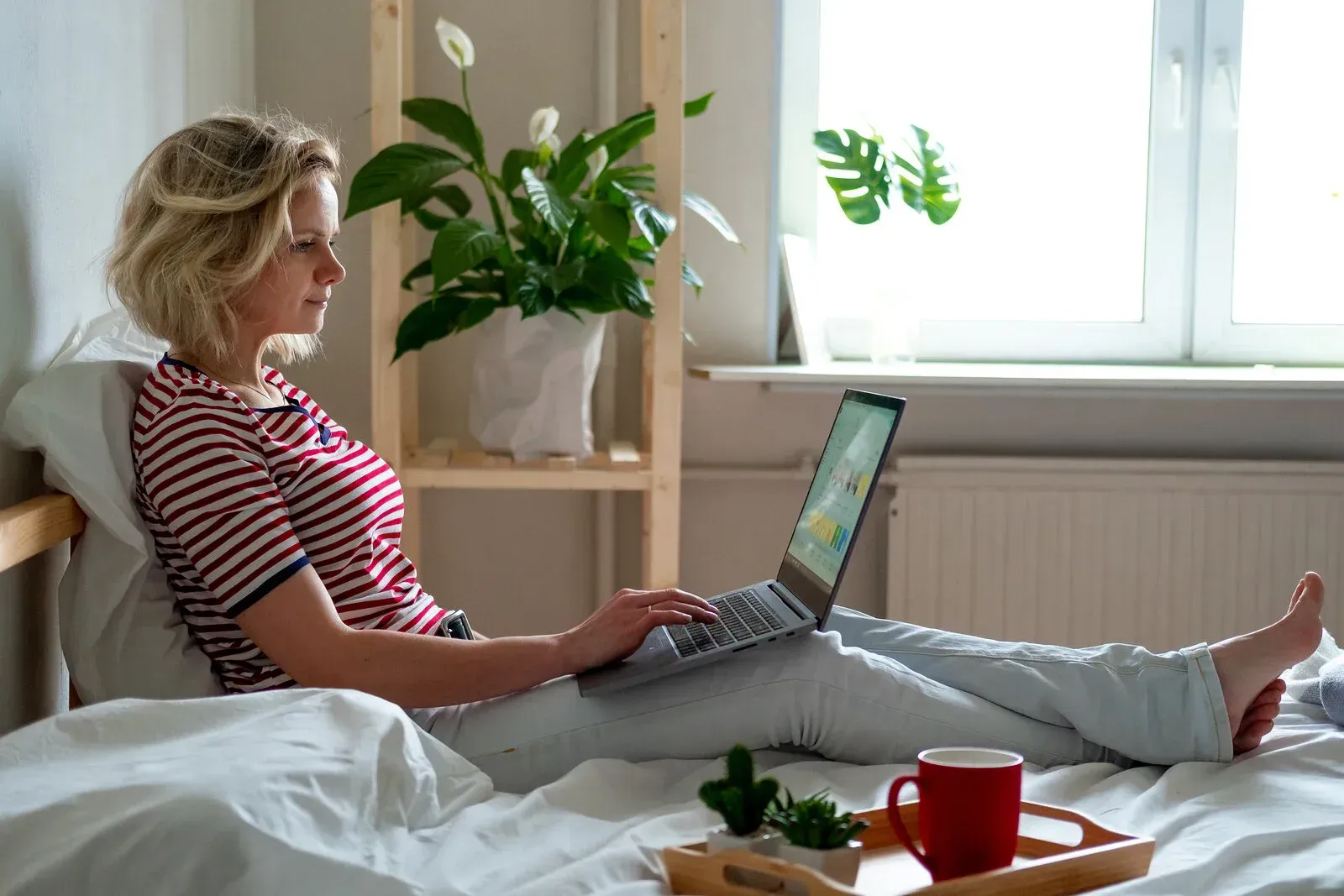 Woman in bed using a laptop for online therapy; red striped shirt, white sheets. Tray with mug and plants nearby.