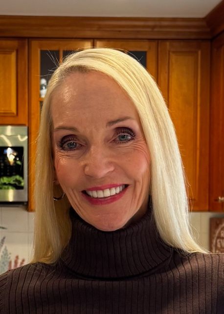 Woman with long blonde hair smiles, wearing a brown turtleneck in a kitchen with wooden cabinets.
