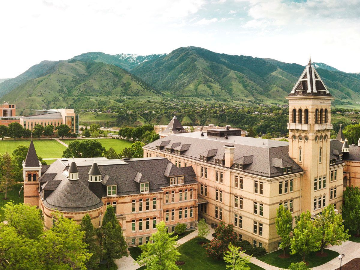 Utah University building in Logan with tower and mountains in the background. Green trees and grass in front.