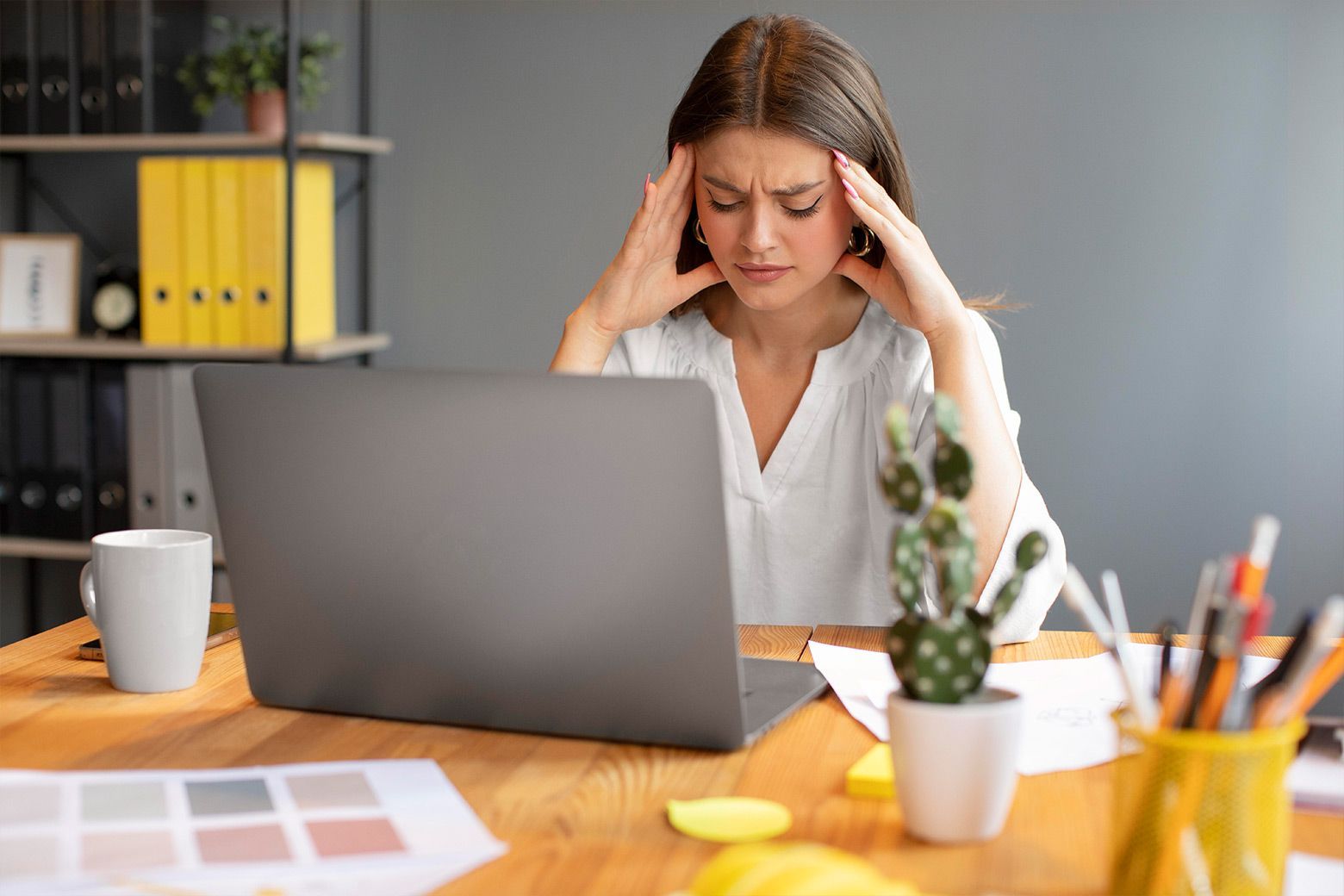 Woman with headache, hands on temples, seated at desk with laptop.