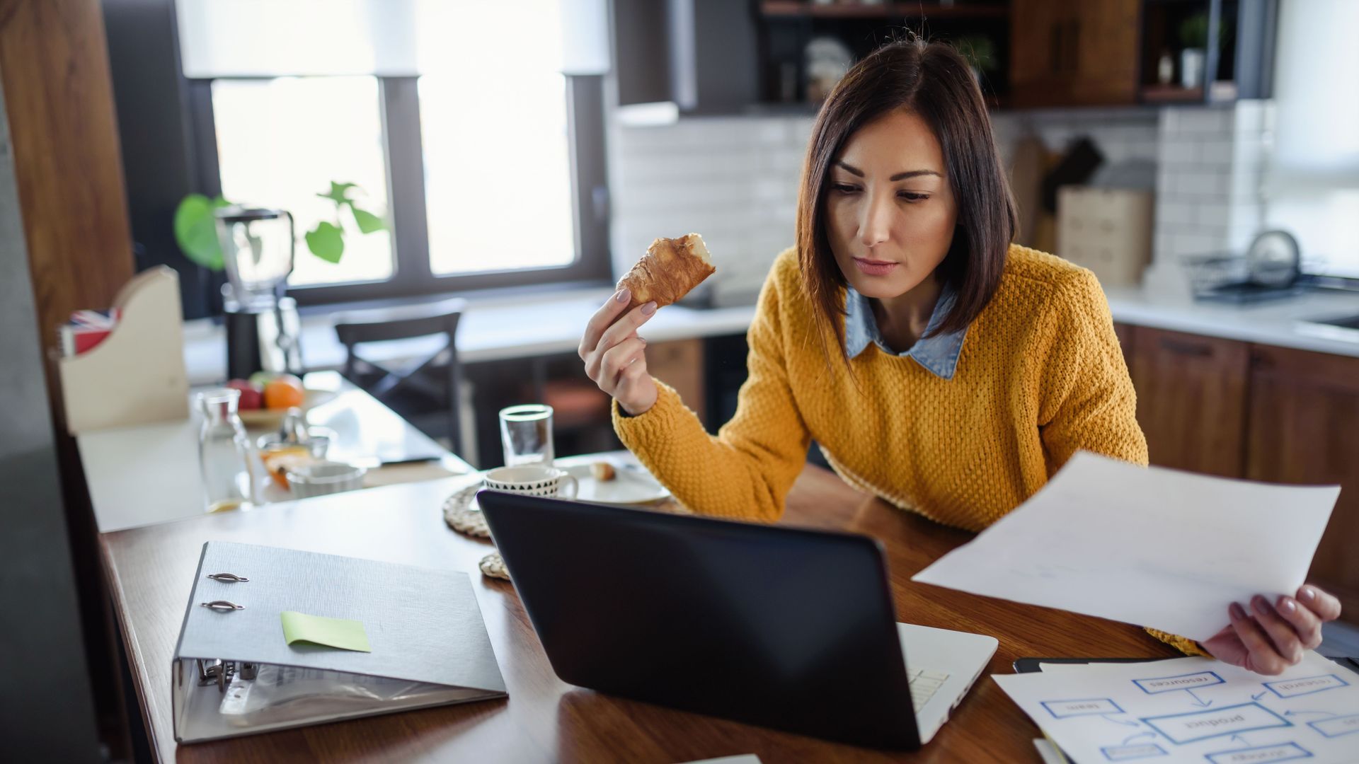 Woman working on laptop at home, holding food and documents in kitchen.