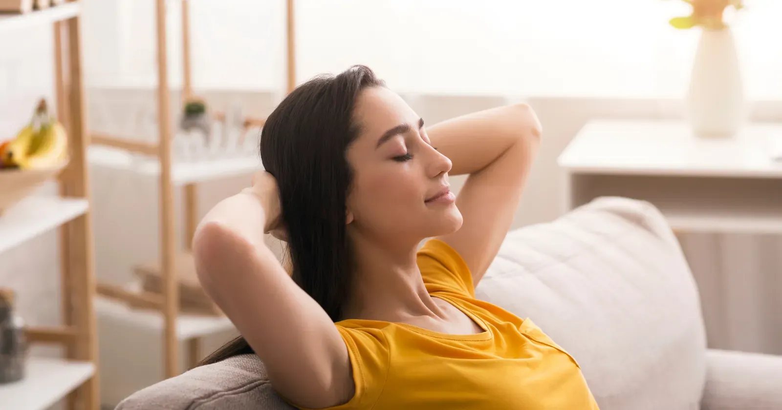 Woman relaxing on a couch with her hands behind her head, eyes closed in a brightly lit room. 