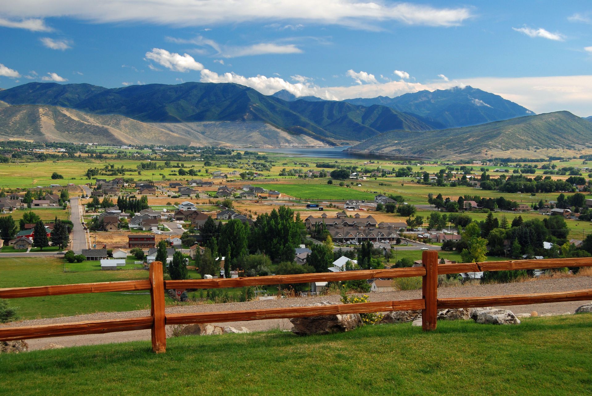 Rural Utah. Rolling green valley, small town, and distant mountains under a blue sky, viewed over a wooden fence.