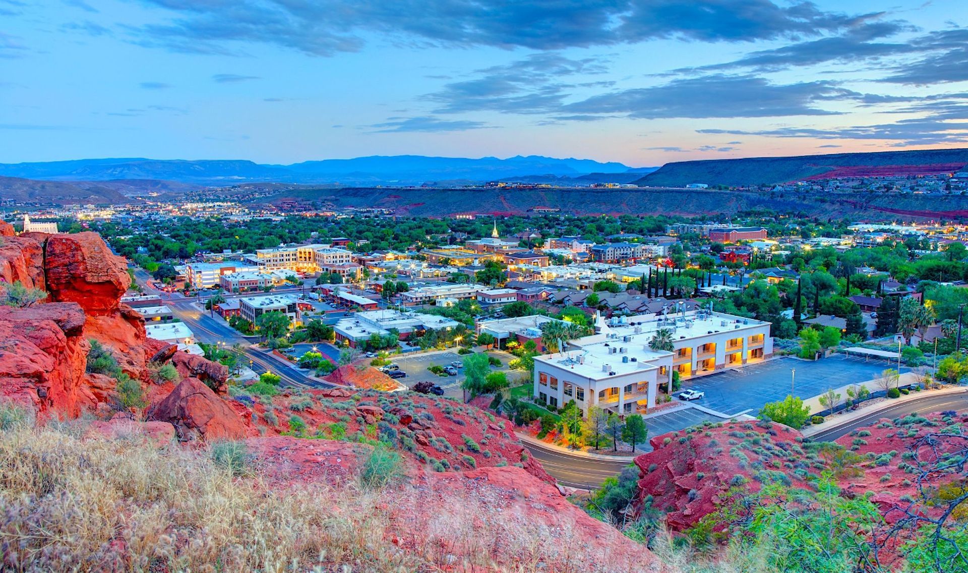 View of St. George, Utah at dusk, with red rock formations in foreground and cityscape stretching to distant mountains.