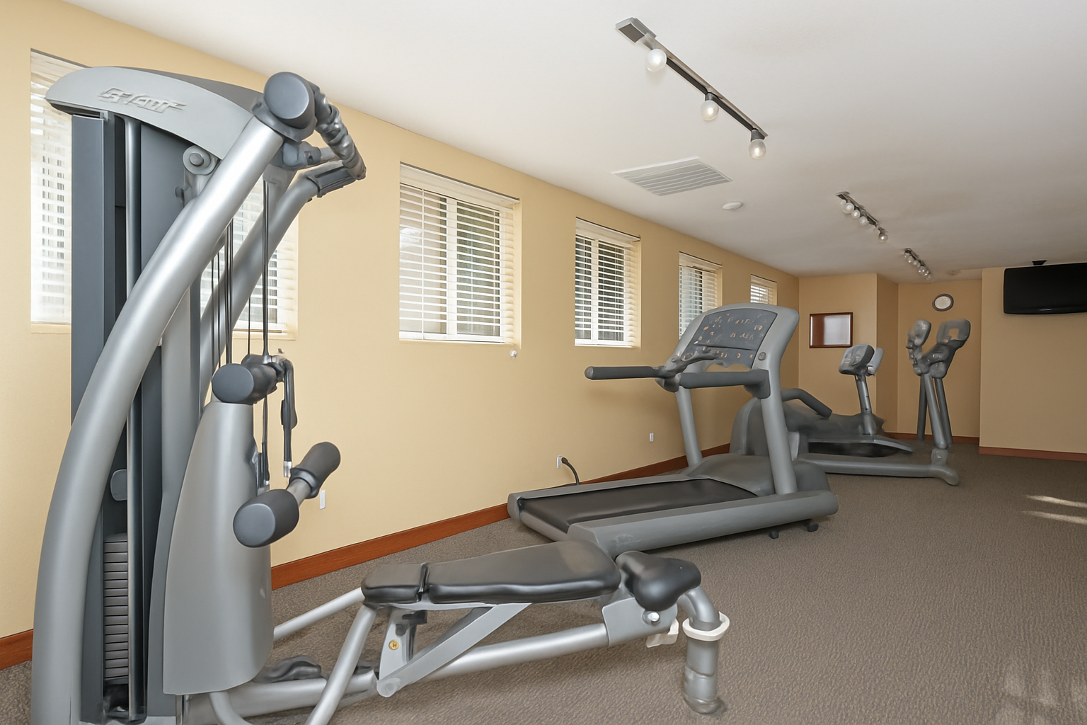 Small gym with elliptical, recumbent bike, TV, and a window. Beige walls, carpeted floor.