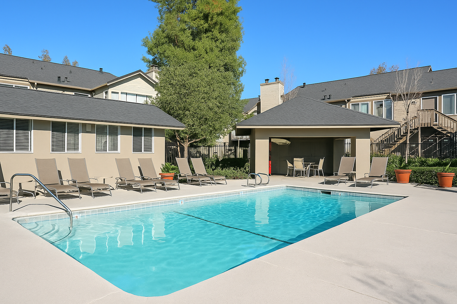 Pool area with lounge chairs, a covered seating area, and apartments in the background on a sunny day.
