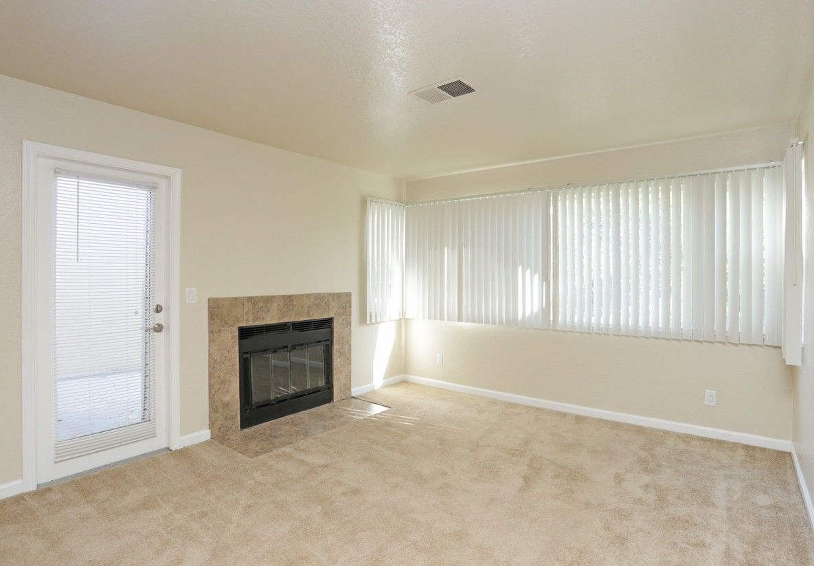 Empty living room with fireplace, door, and large window, beige carpet.
