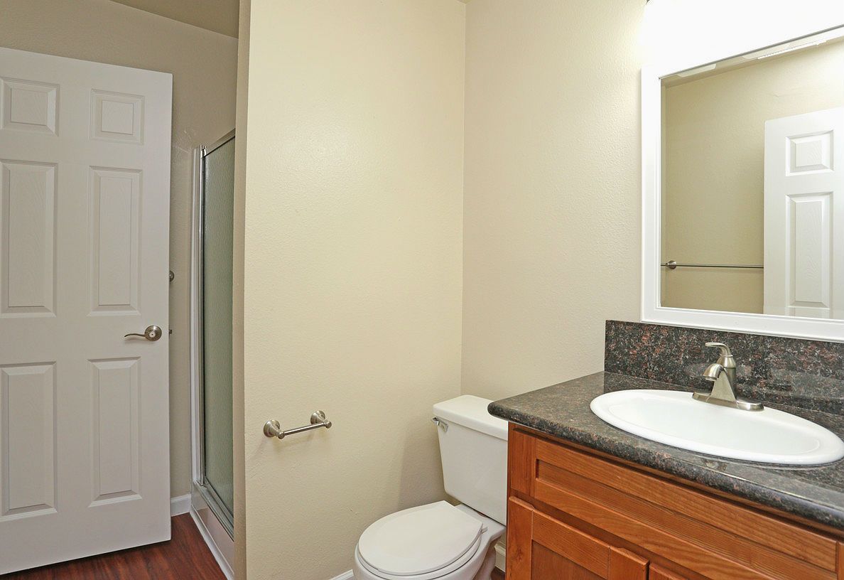 Bathroom with toilet, sink, shower, and closed door. Beige walls, brown wood cabinetry.