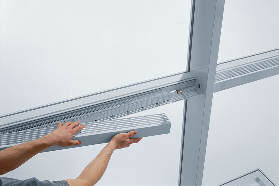 Eine Person montiert eine silberne, lamellenförmige Lüftungsplatte in einen Rahmen an einer weißen Decke.