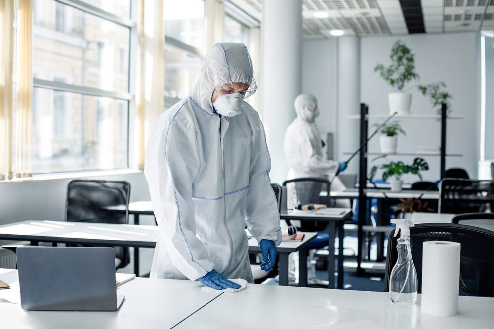 A man in a protective suit is cleaning a desk in an office.