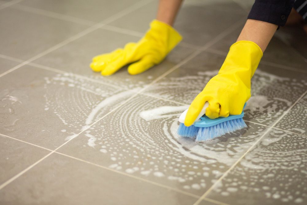 A person wearing yellow gloves is cleaning a tile floor with a brush.