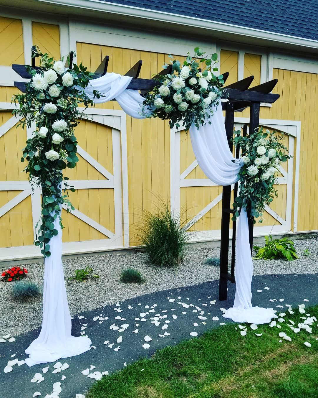 A wooden arch decorated with white flowers is in front of a yellow barn.