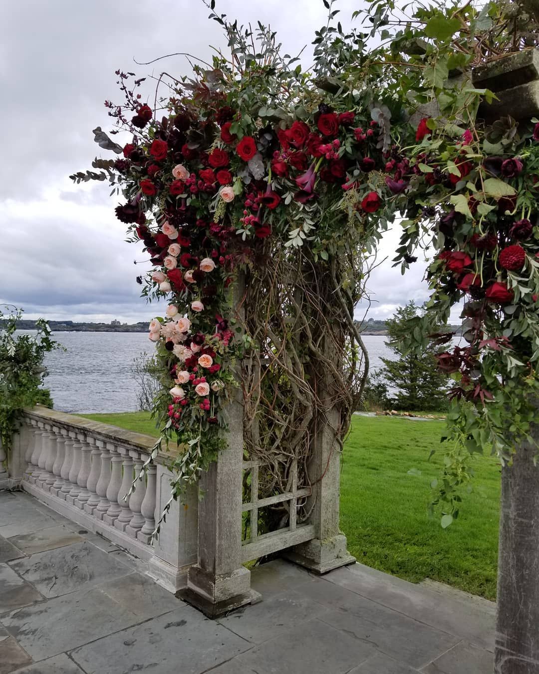 A large arch decorated with red roses and greenery is in front of a body of water.