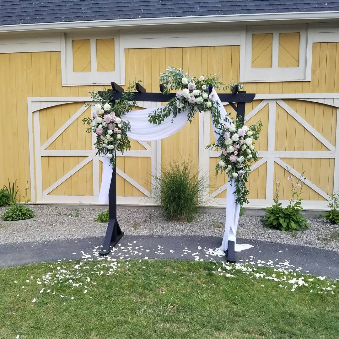 A wooden arch decorated with flowers is in front of a yellow barn.
