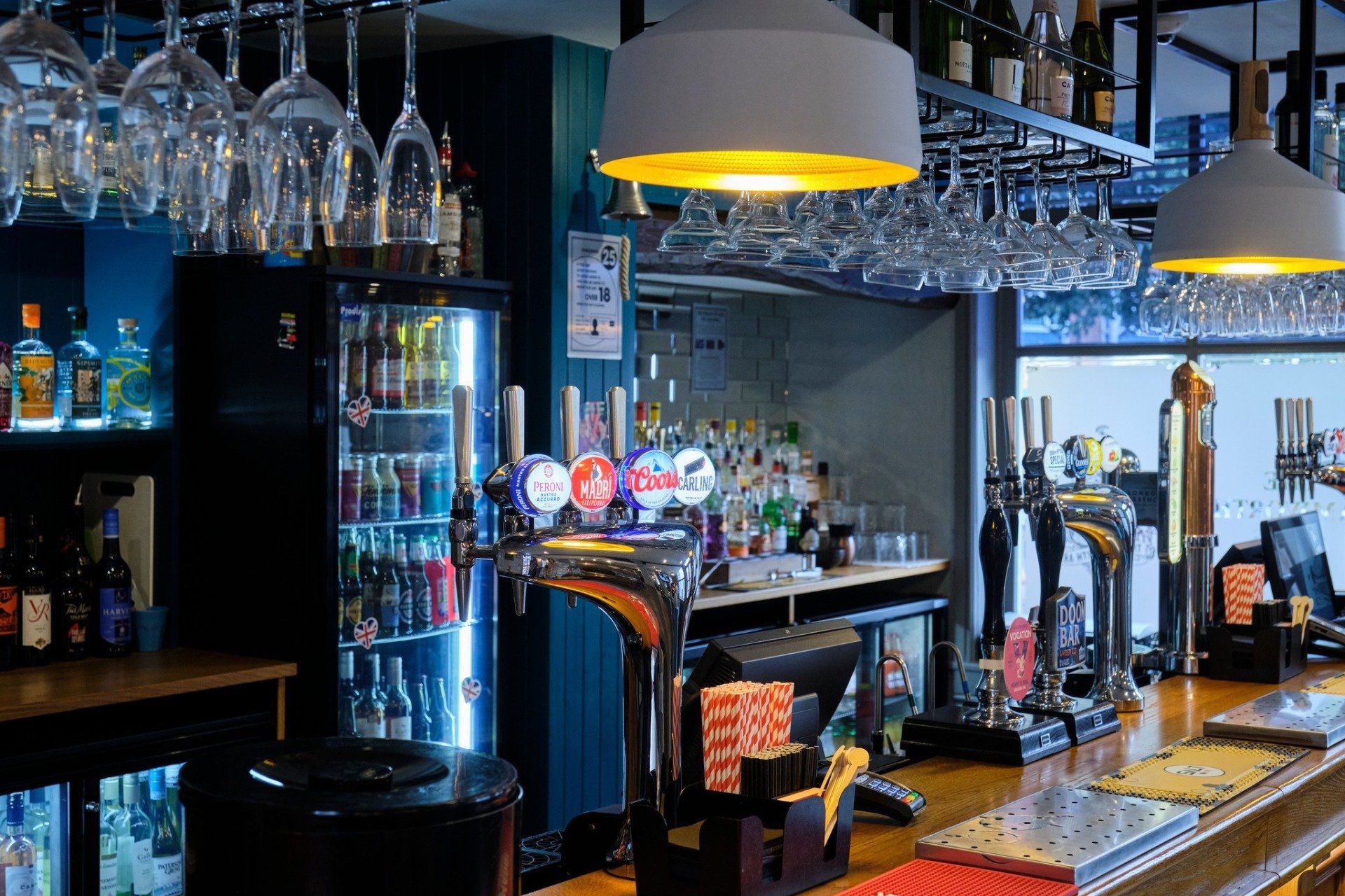 Bar interior with taps, beer fridge, hanging glasses, and overhead lights.