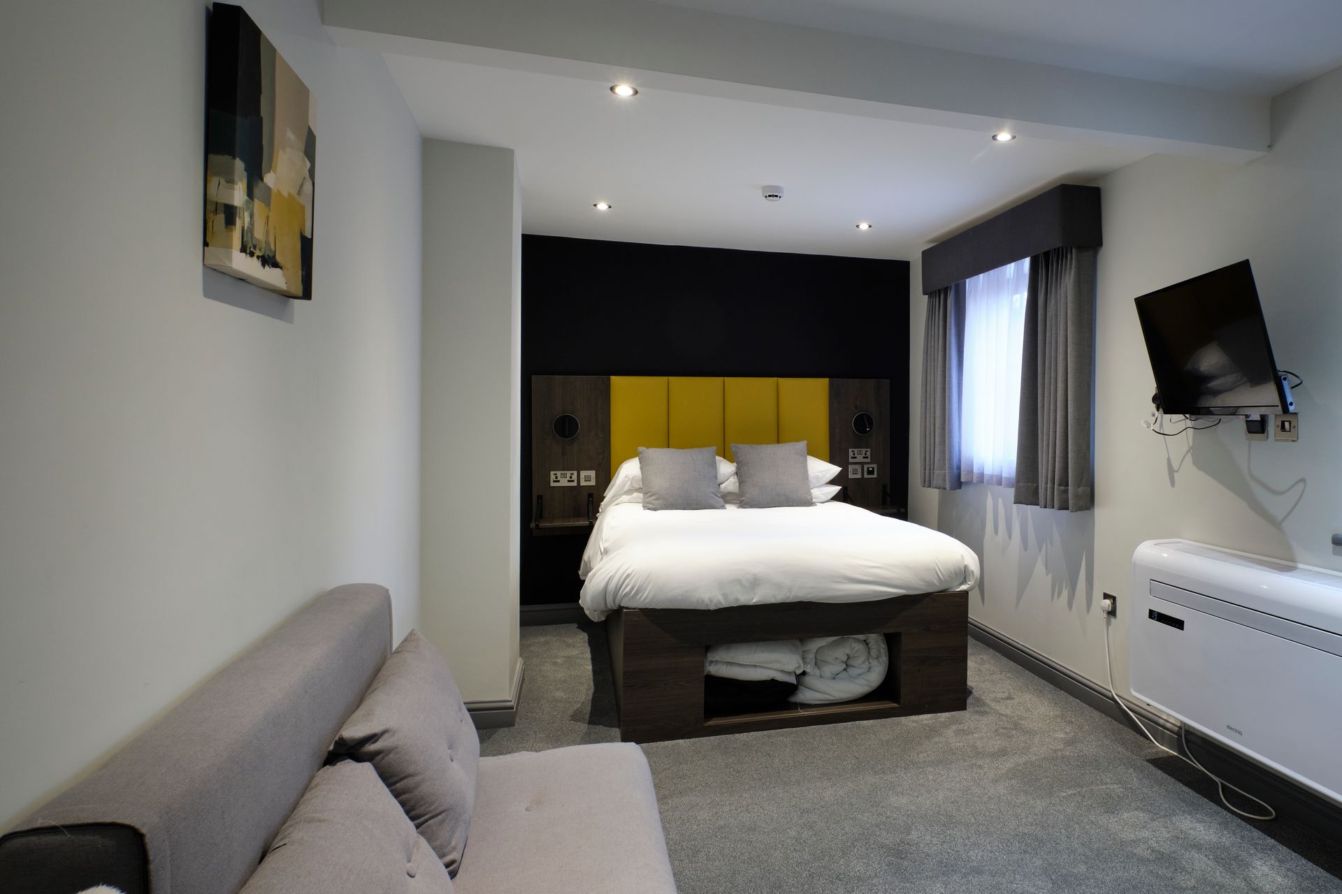 Hotel room with bed, sofa, TV, and window; gray carpet, yellow headboard, black wall.