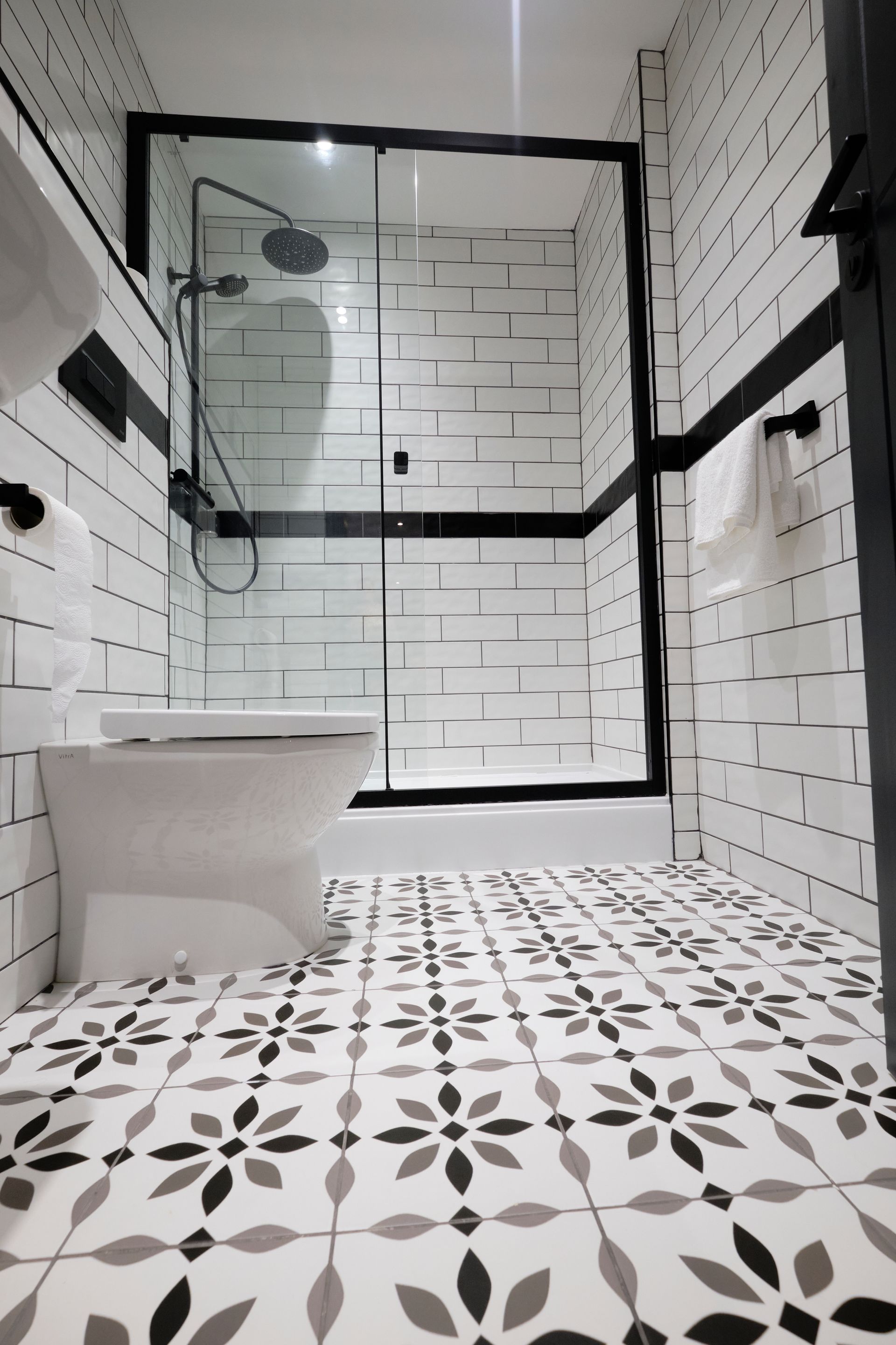 Bathroom with white brick-like tiles, a floral patterned floor, and black trim around the shower and door.