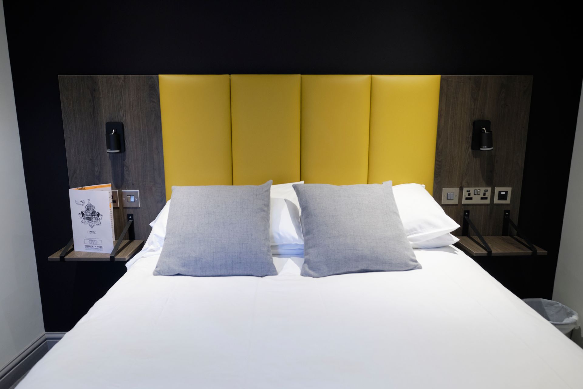 A hotel bed with gray pillows and a yellow headboard, flanked by small nightstands with lamps and outlets.