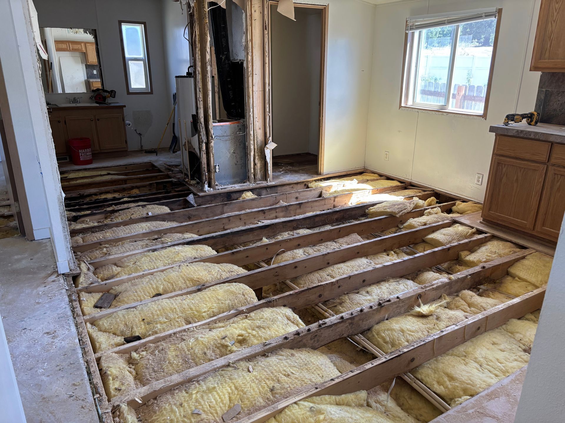 Interior of a home gutted for renovation; exposed floor joists with insulation, damaged walls, and cabinets.