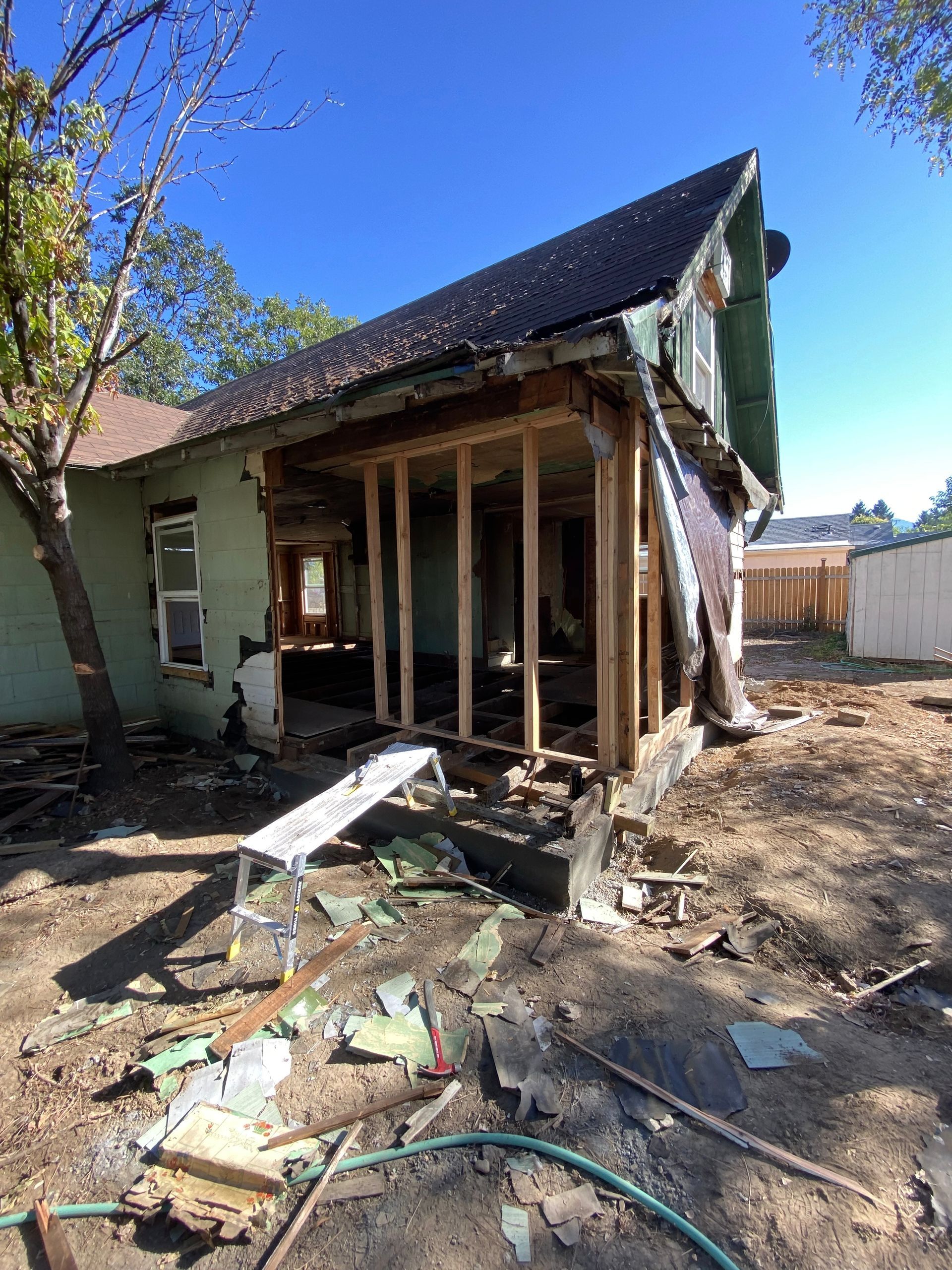 Exterior view of a house under renovation; green siding, exposed framing, debris on the ground, blue sky.