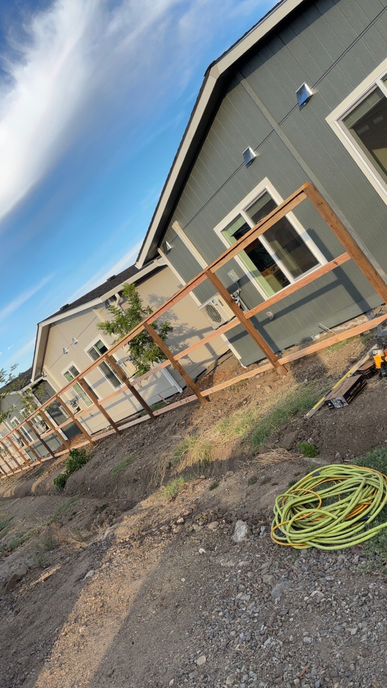 Wooden fence being built between two-tone houses, with a coil of yellow hose on the dirt ground.
