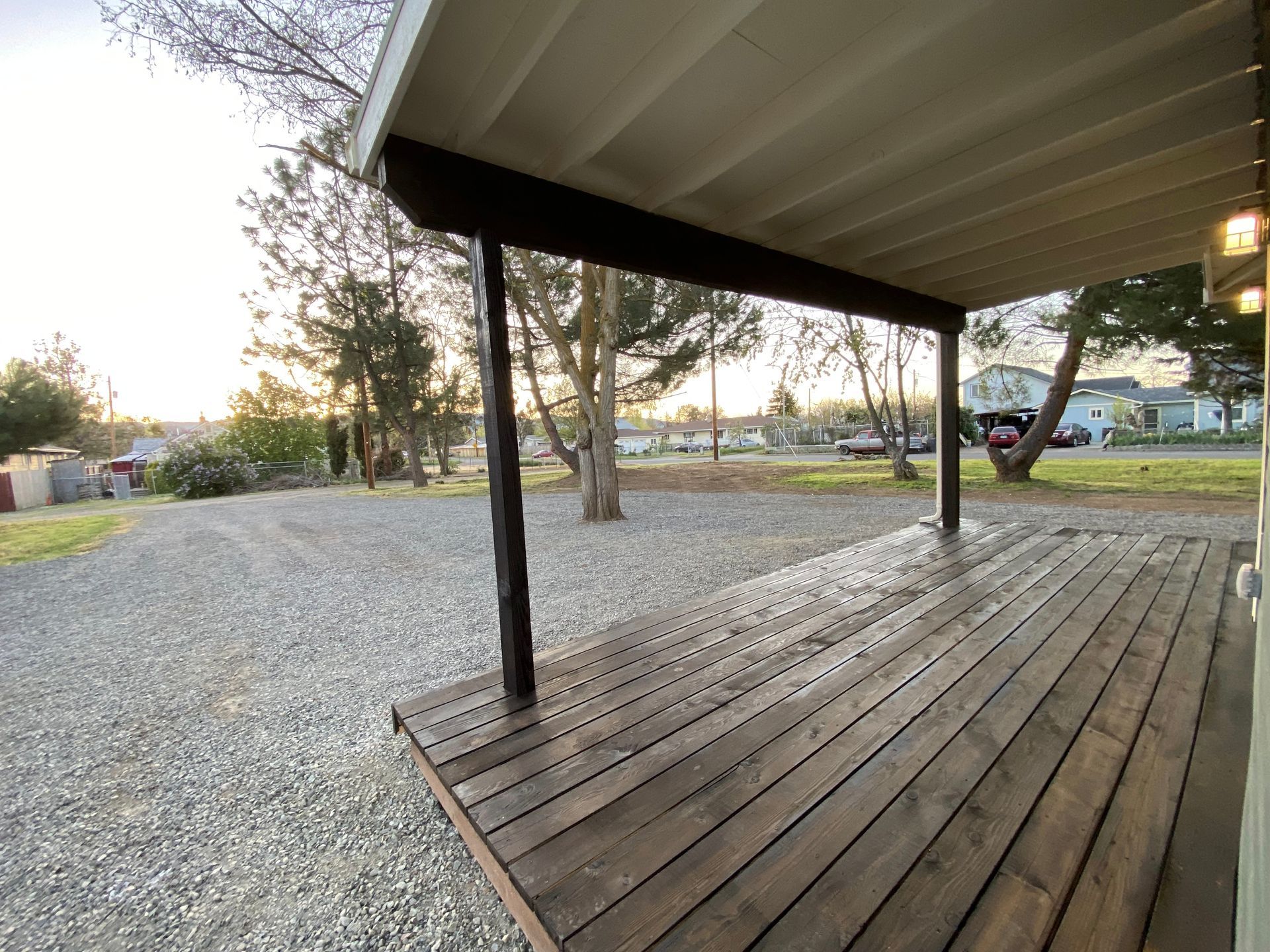 Covered porch with wooden deck overlooking a gravel driveway and trees at dusk.