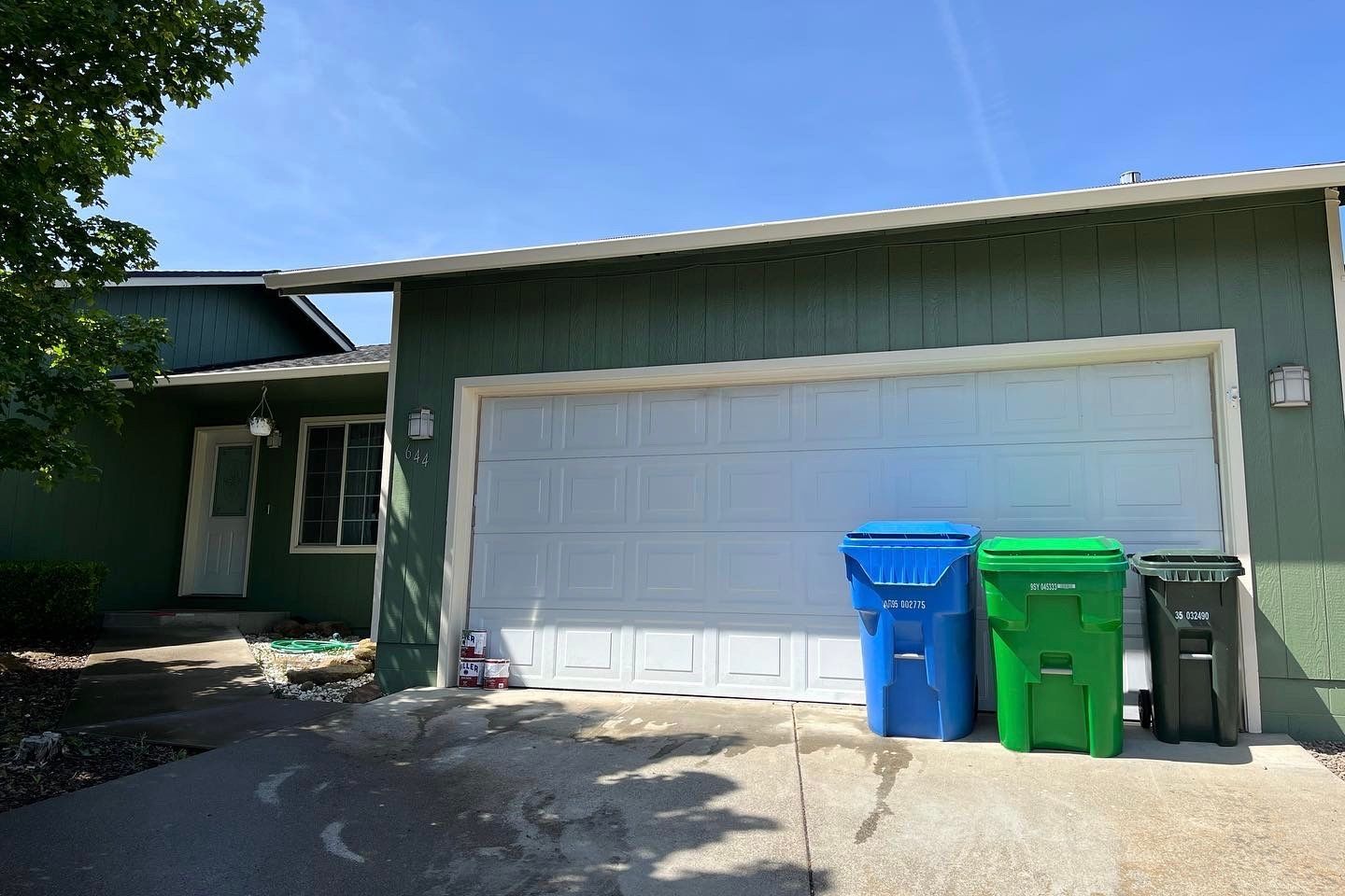 Green house with white garage door; blue, green, and black trash bins in front.
