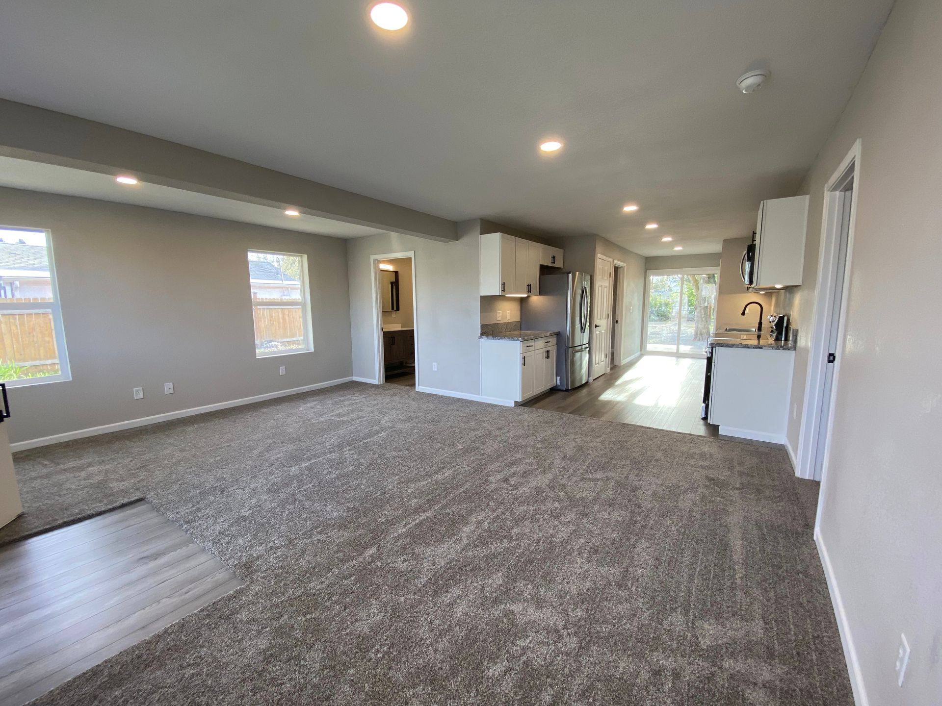 Spacious living area with carpet, light gray walls, and white cabinets. Kitchen and windows are visible.