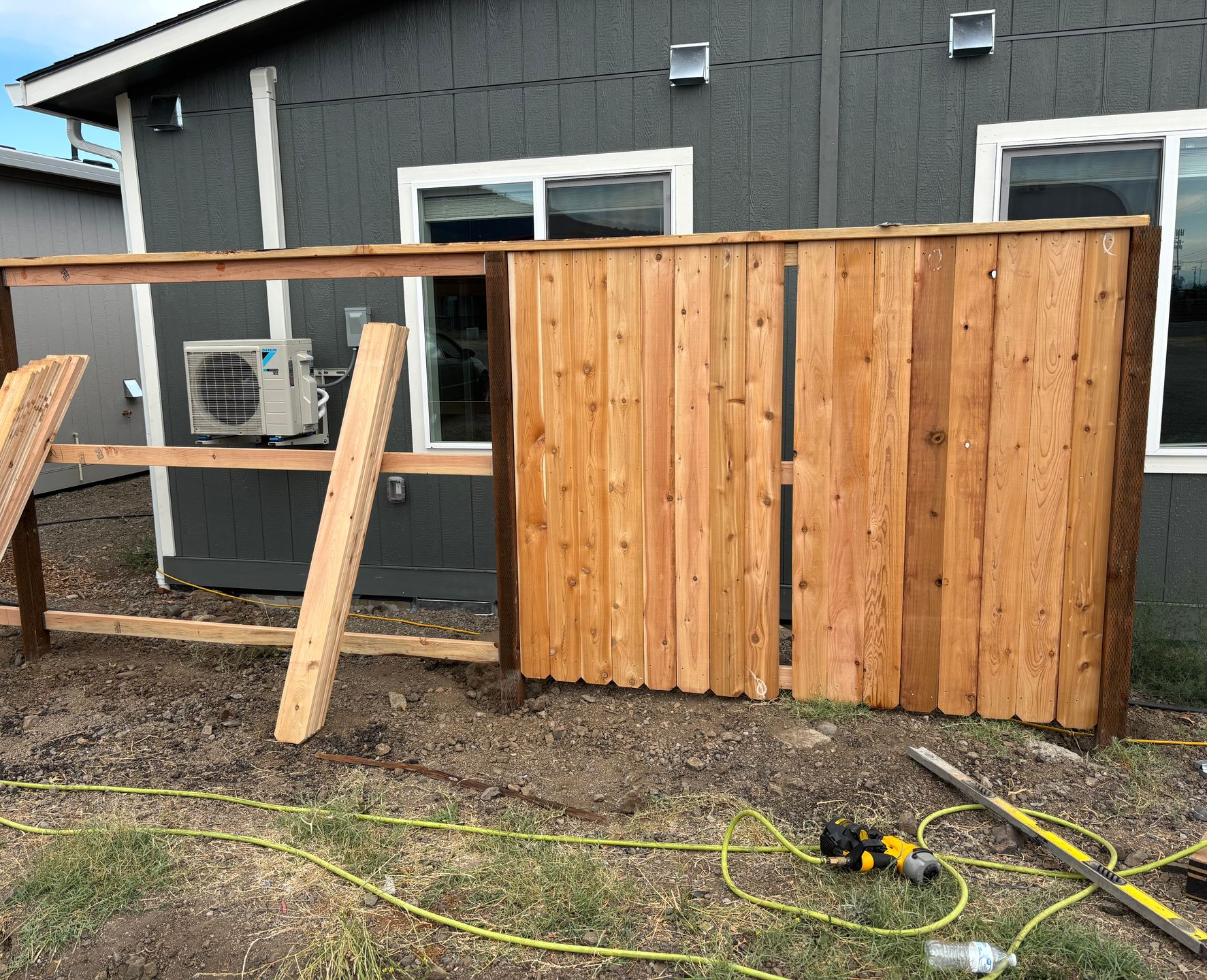 Wooden fence under construction next to a gray house; air conditioner visible.