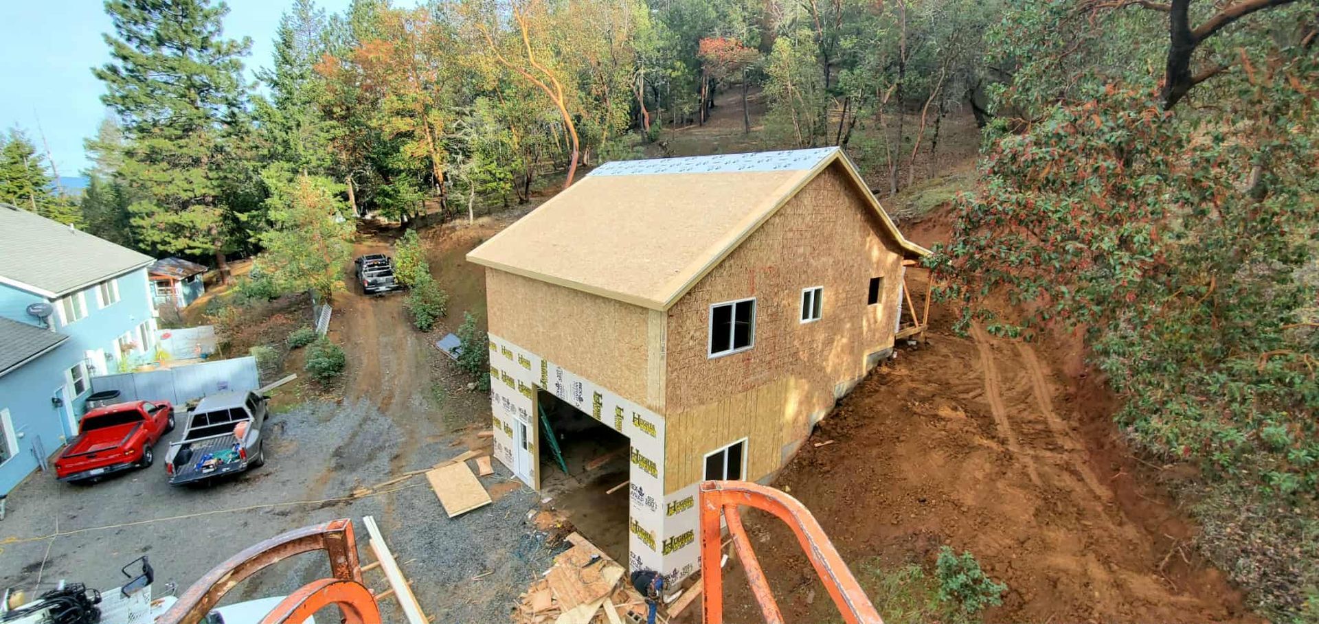 A two-story house under construction on a hillside. Cars and trees are in the background.